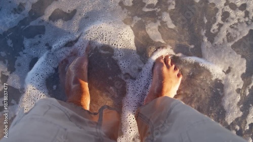 POV view shows bare feet on wet sand as crystal-clear water gently washes over ankles. A peaceful summer vacation moment with soft waves, slow steps, and oceanfront serenity.