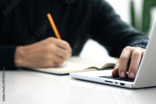Closeup image of a male writing notes in his notebook while using laptop. Boy working on new project