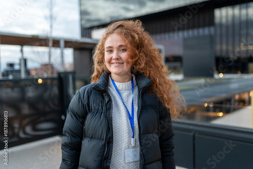 Slika na platnu Young woman smiling, wearing a puffer jacket and blank id badge, standing outdoo