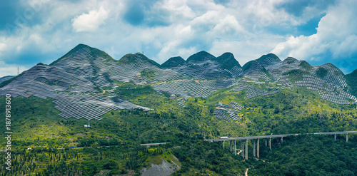 Massive solar panel farm installed on green mountain slopes for renewable energy production under a bright cloudy sky.