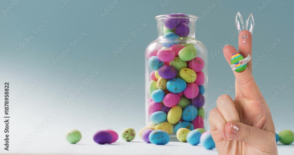 Fototapeta premium Image of fingers with easter bunny and easter egg over jar of easter eggs on blue background