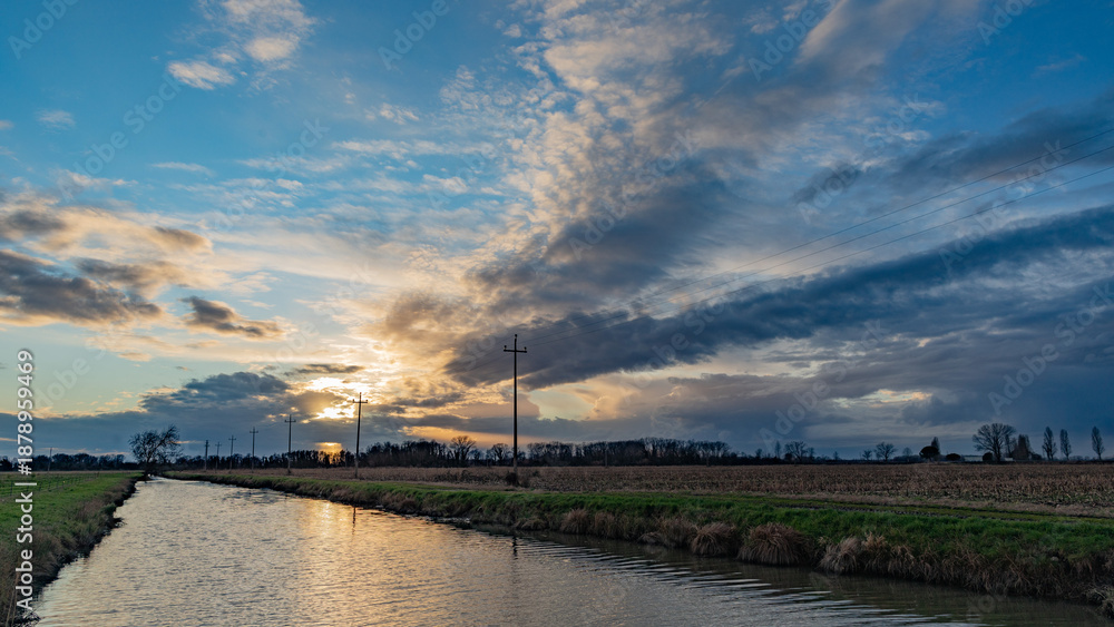 Obraz premium Vista panoramica su di un ambiente naturale di campagna in pianura nel nord Italia, con campi ed un canale pieno d'acqua, al tramonto, col cielo a metà tra sereno e nuvoloso