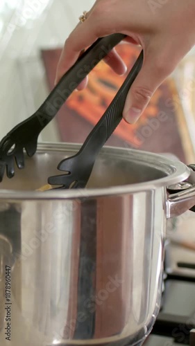 Vertical video: Lowering hand with black tongs placing fresh pasta into steel pot on stove, to cook