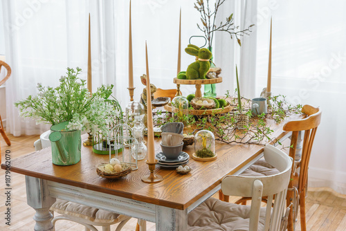 Stylish Easter table decor with candles, greenery, bunny figurines and natural elements in bright spring room interior. Cozy festive dining setup with wooden table, soft daylight and seasonal details