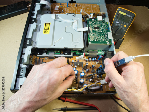 Photos Close-up of technician's hands using a multimeter to diagnose or repair complex electronic circuitry inside an open DVD/Blu-ray player or similar device