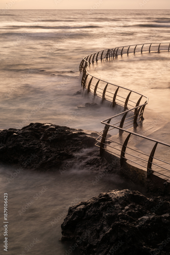 Fototapeta premium A rocky coastline with a metal fence and a pier at La Punta del Hidalgo in Tenerife, Canary Islands. The water is choppy and the sky is cloudy