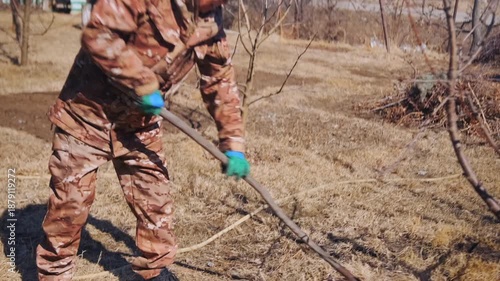Man Cleans Yard - Worker Clearing Yard Debris Energetically - Person Gathering And Raking Fallen Branches And Leaves Diligently - Man Dressed In Camouflage Working Hard To Tidy Garden Area