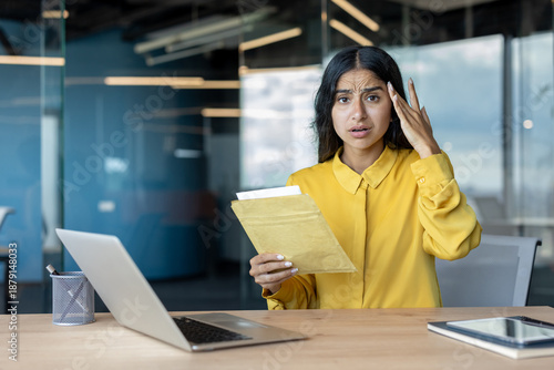 Fotografía Portrait of a worried young Indian woman sitting at a desk in the office, holdin