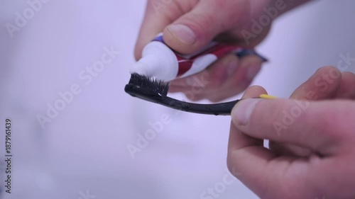 Toothbrush receiving white paste carefully, Gentle squeezing of small toothpaste tube for brushing, Detailed view of hands carefully dispensing toothpaste onto toothbrush in bathroom environment