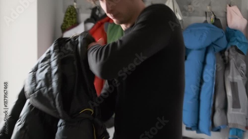 caucasian man hallway choosing winter coat, tying headband, sliding on puffer jacket, checking pockets, grabbing backpack, colorful coats on hooks, morning light, focused casual commute prep