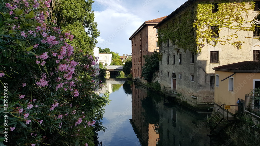 Αυτοκόλλητο Picturesque canal in Portogruaro, Italy: calm waters mirror ivy-covered brick buildings and stone bridge under blue skies