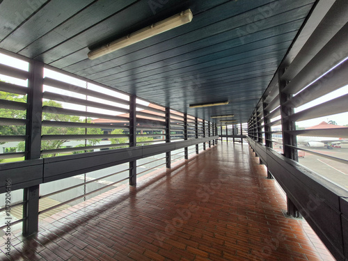 Classic airport terminal connecting corridor with wooden ceiling, ventilated wooden walls, and brown tile floor, designed for natural airflow, comfort, and traditional architectural style.