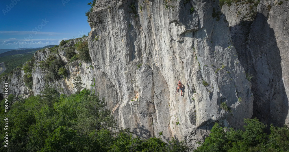 Fototapeta premium AERIAL: Rock climber scales a tall limestone face in the famous Osp climbing area. Shirtless man high above the forest canopy, set against a backdrop of beautiful Karst topography and bright blue sky.