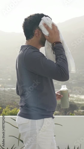 Vertical video: Sun heating man wiping forehead with white towel on balcony, holding green bottle