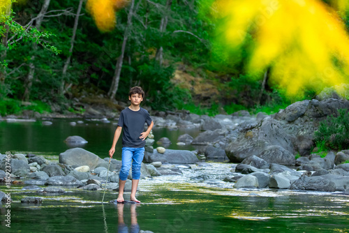 Teenager boy is standing on a rocks in the mountain rocky river with clear green waters in summer forest. Scene under branches with green lush foliage. Sooke potholes park, Vancouver island, Canada.
