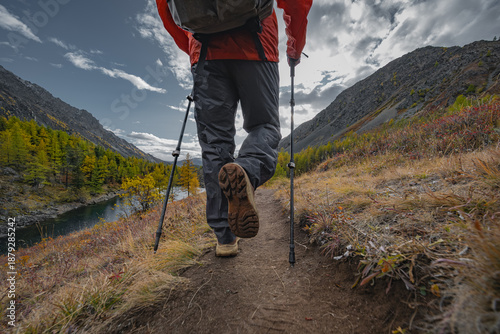 A hiker in bright gear with poles and backpack walks a forested mountain road overlooking an autumn lake. Low rear-angle shot captures boot sole and movement under a cloudy sky.
