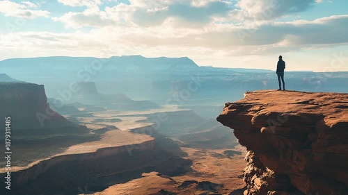 Man stands on cliff overlooking vast canyon landscape at sunset.