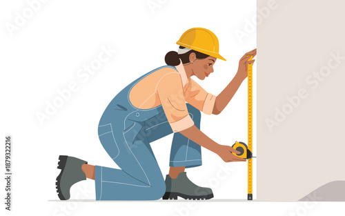 A woman in overalls and hard hat measuring a wall with a tape measure, kneeling on one knee, in a construction setting.
