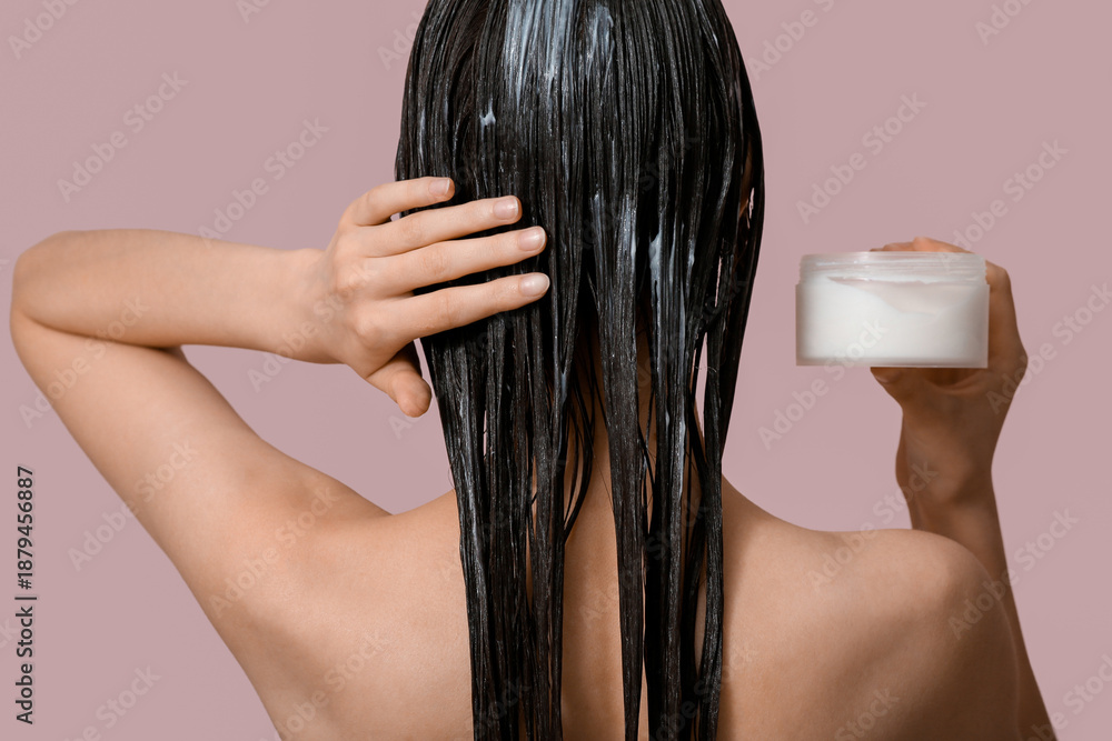 Fototapeta premium Beautiful young woman with jar of cosmetic product applying hair mask on pink background, back view