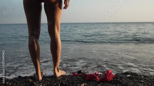 Woman sexy legs bikini undressing on black pebble beach near gentle sea waves during vacation