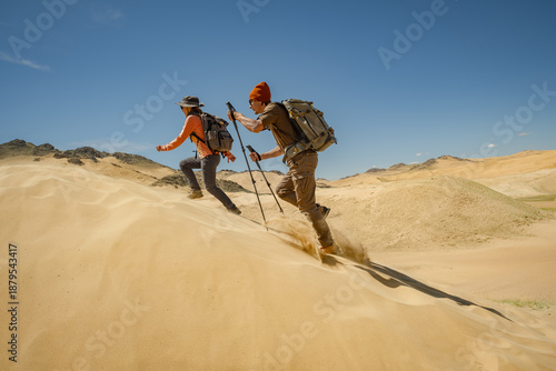 Foto Two hikers with backpacks and trekking poles running or climbing up a steep sand dune under a bright blue sky in an arid desert landscape