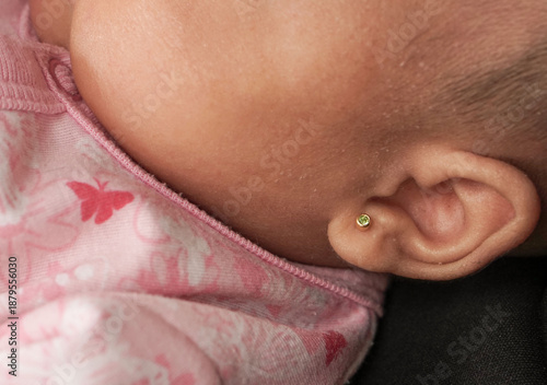 Extreme close-up shot focusing on the delicate ear of an infant wearing a small, golden stud earring with a green stone. The baby's skin texture is visible, resting against soft pink fabric patterned