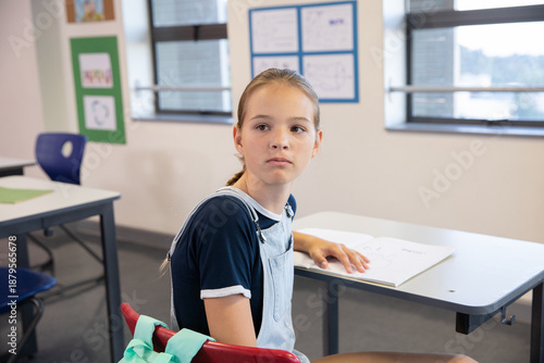 Adolescent student sitting at desk reviewing open workbook at school with red chair and jacket