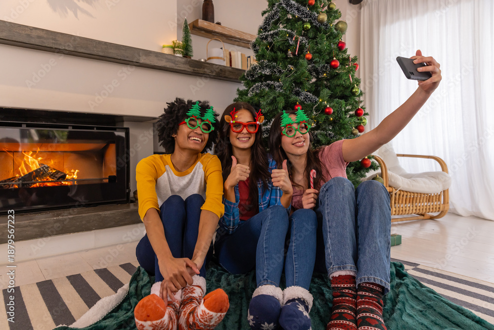 Obraz premium Diverse female friends sitting at home by christmas tree in novelty glasses holding smartphone