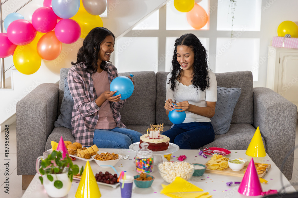 Obraz premium Diverse mom and teenage daughter inflating bright blue balloons in living room with birthday cake