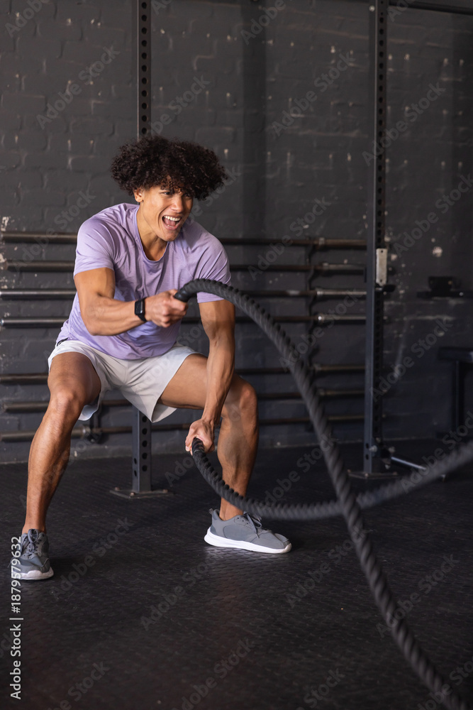 Fototapeta premium African American man wearing lavender shirt and gray shorts swinging battle ropes in gym studio