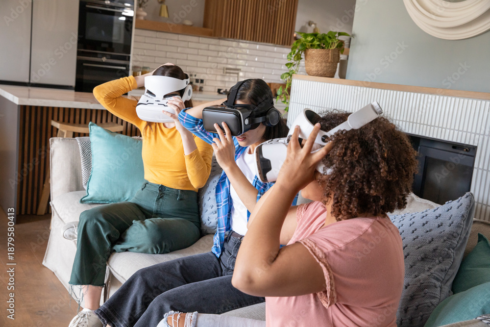 Fototapeta premium Diverse female friends wearing VR headsets while sitting on sofa among patterned cushions at home