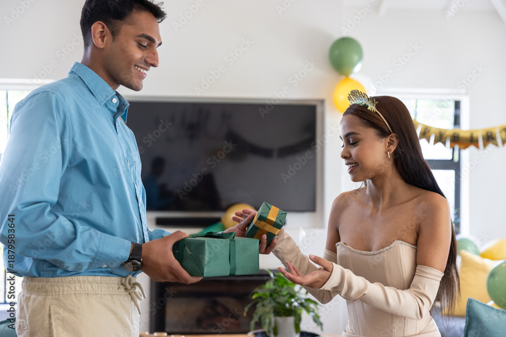Naklejka premium Diverse couple exchanging green-wrapped gift at party in living room with balloons, banner
