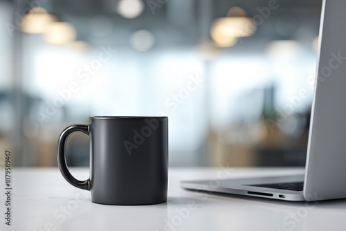A black ceramic mug stands on a white table next to a silver laptop against a blurry background of a modern office