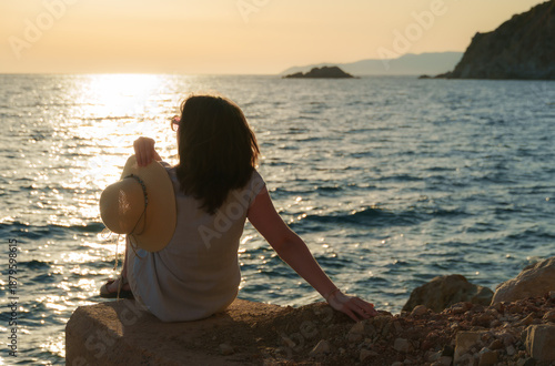 Young woman in sunglasses relaxing on the rocky coast at sunset, looking at the water with a straw hat in hand during summer vacation.