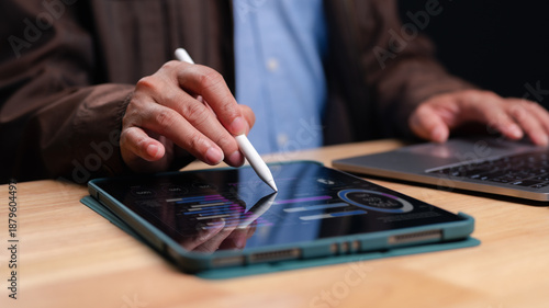 Professional businessman analyzes financial data reports using digital tablet stylus. Modern office workspace features laptop computer wooden desk surface. Detailed charts graphs display market
