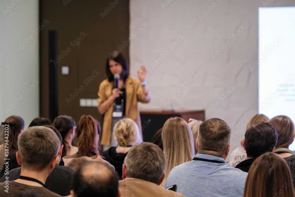 Obraz premium Bright speaker presents at conference. Audience listens attentively. Woman holds microphone, raises hand. Modern meeting space with projector screen. 