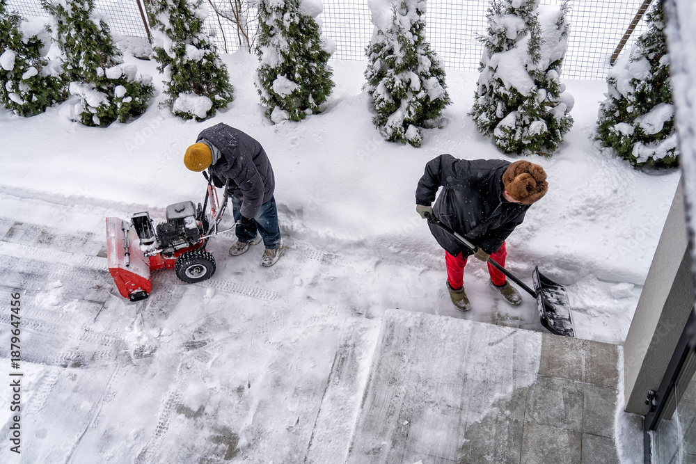 Fototapeta premium Two people diligently clearing heavy winter snow from residential driveway and walkway, one operating powerful snow blower and other shoveling by hand, showcasing essential cold weather maintenance an