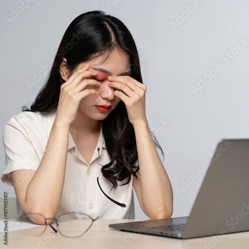 Stressed Asian woman with tired eyes rubbing them while working on laptop.
