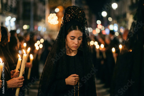 Procesión nocturna en primer plano, mujer vestida de mantilla negra con guantes de encaje sosteniendo un rosario