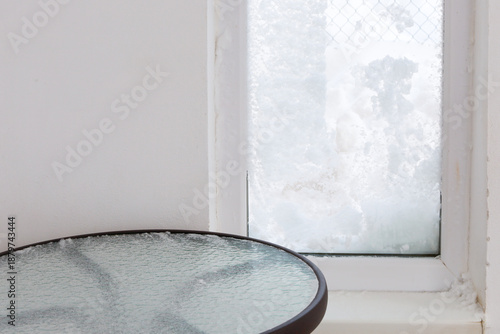 A snow-covered window and a glass table topped with a thin layer of ice convey the chilly atmosphere of a winter day indoors. Light wall tones and natural light filtering through the frost-covered gla