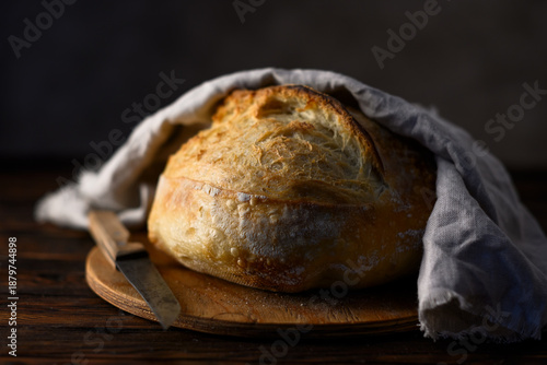 Sourdough bread with cracked crust and rugged texture wrapped in a linen towel on a rustic wooden table with knife. Artisan farmhouse-style loaf close up