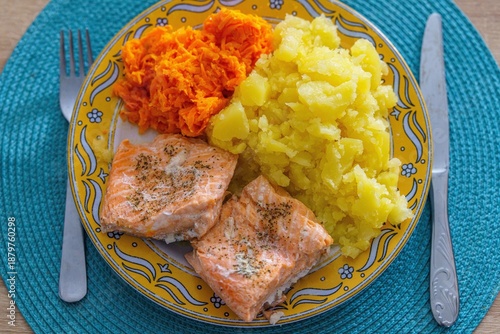 Zdrowy, domowy obiad składający się z pieczonego fileta z łososia, gotowanych ziemniaków oraz surówki z tartej marchewki. Danie serwowane na dekoracyjnym żółtym talerzu ze sztućcami na niebieskiej pod