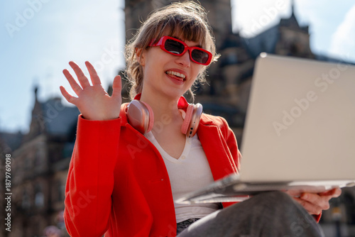 Fotografía Young woman waves while working on laptop outdoors in city during sunny day