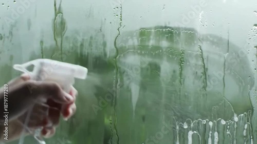 Hand spraying cleaning fluid from a spray bottle onto glass and wiping condensation from a green glass surface. Water droplets and moisture remain on the fogged window