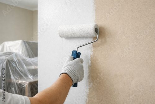 Painting a beige wall white using a roller brush by a gloved hand, with covered furniture in the background, showing home renovation, interior improvement, and fresh wall coating.