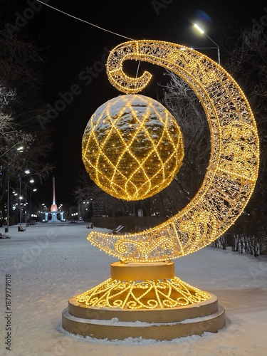 Glowing Christmas installation of a golden crescent moon and sphere in a snowy park at night.