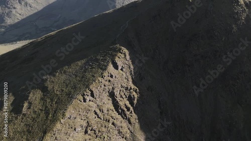 Sunlight hits the side of a tall mountain. A narrow trail is visible near the peak. It is early in the morning.