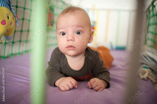 A baby, lying on their stomach in a crib with a purple sheet, looks up with wide eyes, showing curiosity or surprise at something happening during daytime