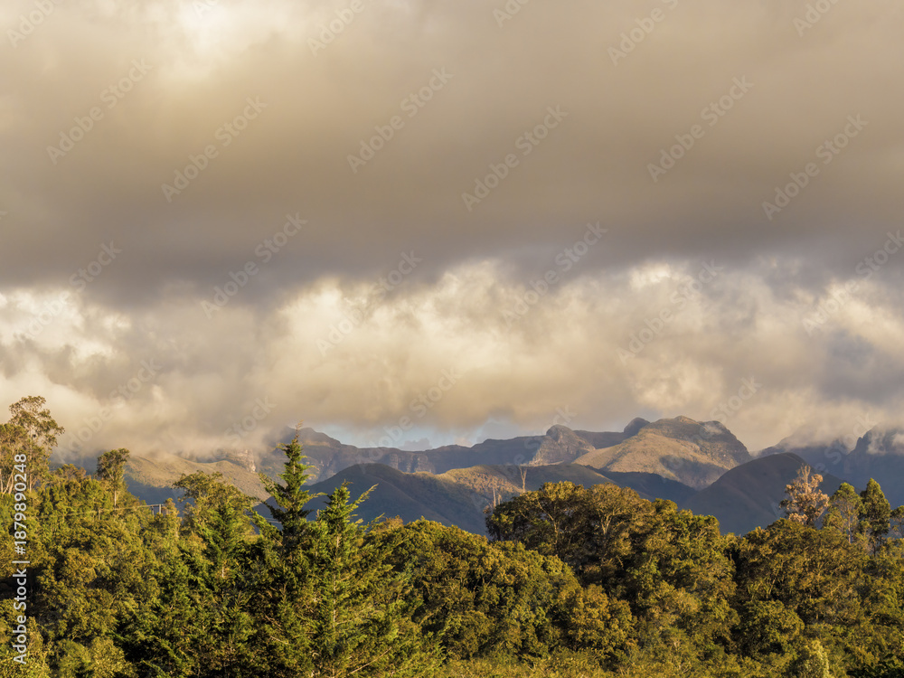 Fototapeta premium The Iguaque natural reserve behind the forest illuminated by the light of sunset, under an overcast sky, at the eastern Andean mountains of central Colombia.
