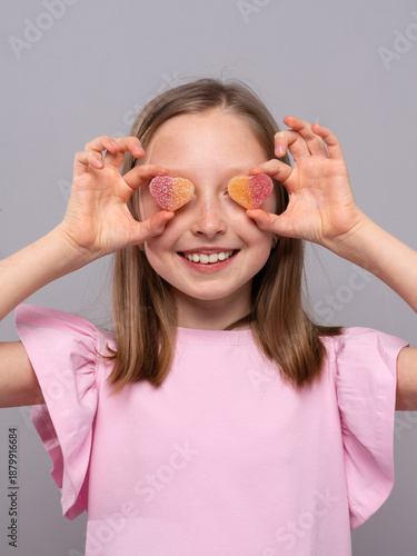 smiling young girl standing against a light gray studio background. She is wearing a soft pink dress and holding two heart-shaped, sugar-coated gummy candies over her eyes like playful glasses. The im